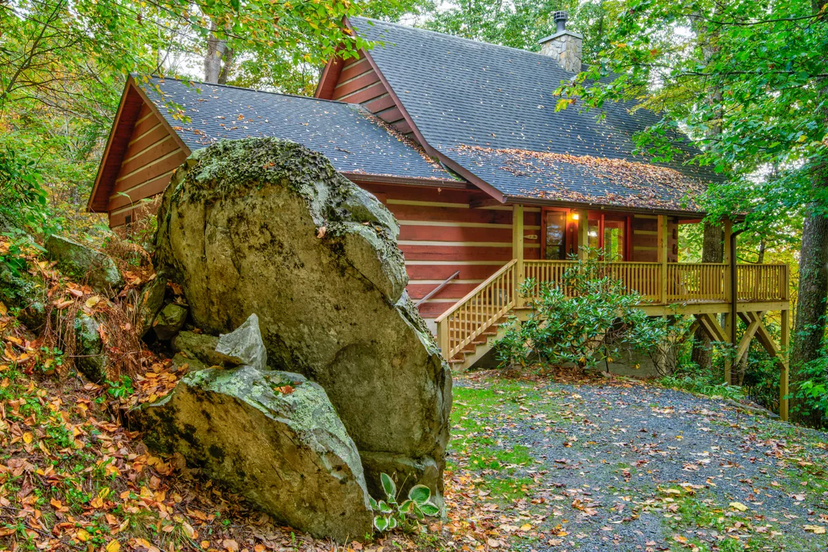 The foreground is dominated by a large boulder, with a charming wooden porch and a distinctive stone chimney visible on the cabin. The scene is surrounded by trees, evoking a sense of adventure and offering a glimpse of the natural landscape couples can explore hand-in-hand just outside the door.