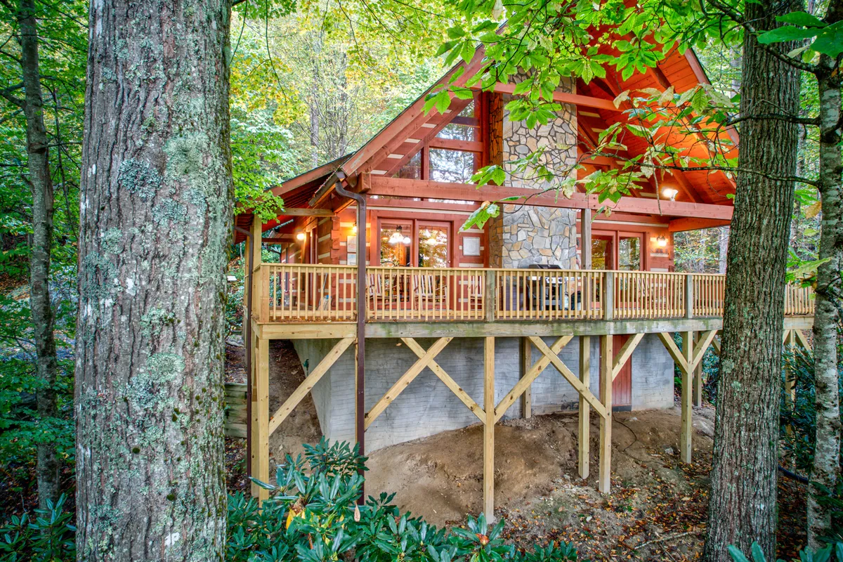 A wide exterior perspective featuring a wooden deck, stone chimney, large windows, and vibrant surrounding foliage. The deck is clearly designed for outdoor gatherings, barbecues, and relaxing in nature, with sunset views and space for evening fires under the stars.