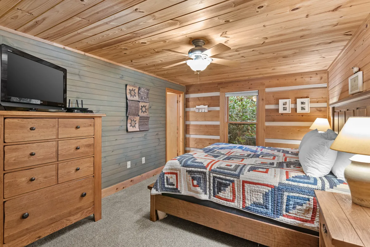 A warm, inviting bedroom with a quilted bedspread, wooden dresser, television, and a window letting in ample natural light. The cozy bedding and patchwork quilt evoke comfort and relaxation. The window offers a glimpse of nature outside, creating a serene environment for rest and rejuvenation.