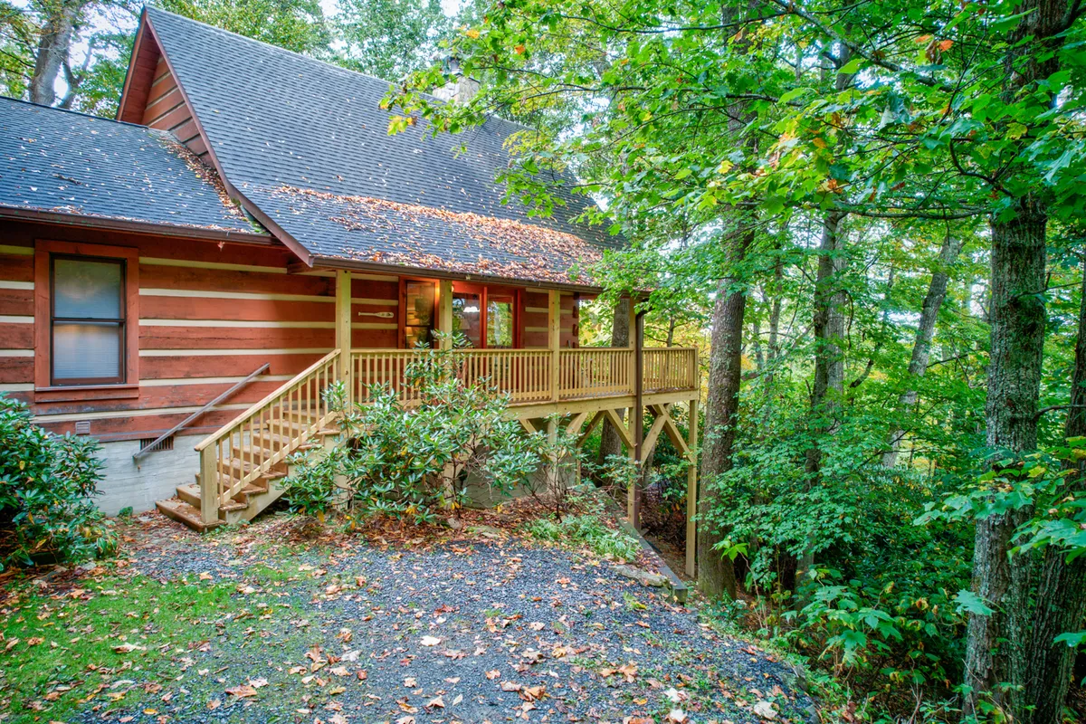 A classic log cabin with a welcoming wooden deck sits nestled among mature trees and lush green landscape. The scene exudes serenity, with the deck offering a vantage point to enjoy the mountain views and ever-changing seasons. The space is ideal for relaxing, gathering, and immersing in the beauty of the outdoors.