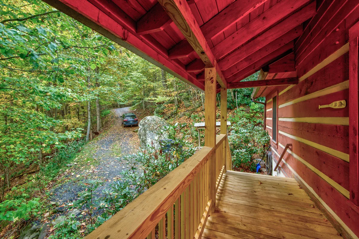 A welcoming cabin exterior with a wooden porch railing and red siding, set beside a pathway that winds into the lush woods. The scene highlights the transition from comfortable shelter to untamed nature, with tall trees framing the cabin.