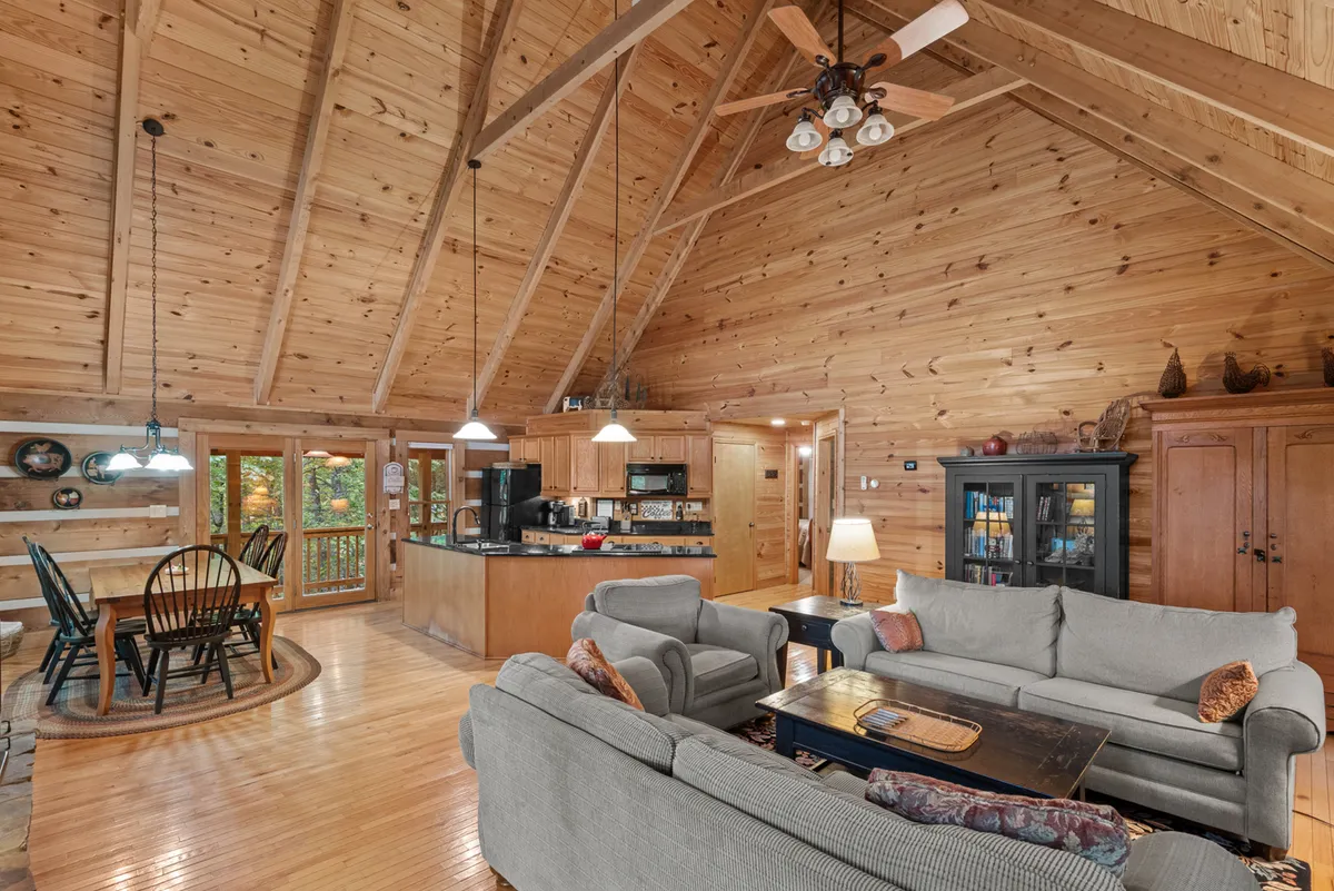 A spacious open-concept living area featuring a high vaulted wooden ceiling, comfortable sofas, a dining table with black chairs seating six, and a modern kitchen area complete with appliances. Large windows offer views of lush greenery outside, and the warm decor creates an inviting, communal atmosphere.