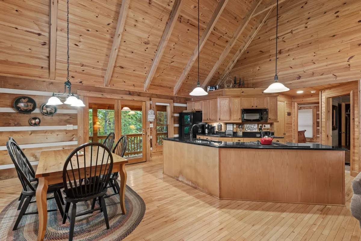 A cozy kitchen and dining area with a kitchen island, a sturdy wooden dining table, and large windows that flood the space with natural light. The arrangement is ideal for family meals and socializing, with direct sightlines between the kitchen and dining spaces.