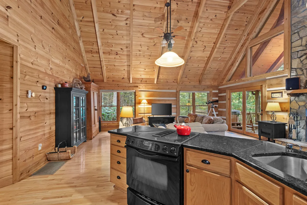 A beautifully styled living area with a kitchen island, comfortable seating, stone fireplace, wooden beams, and large windows letting in natural light. The setting feels communal and stylish—perfect for gatherings or peaceful moments taking in the views.