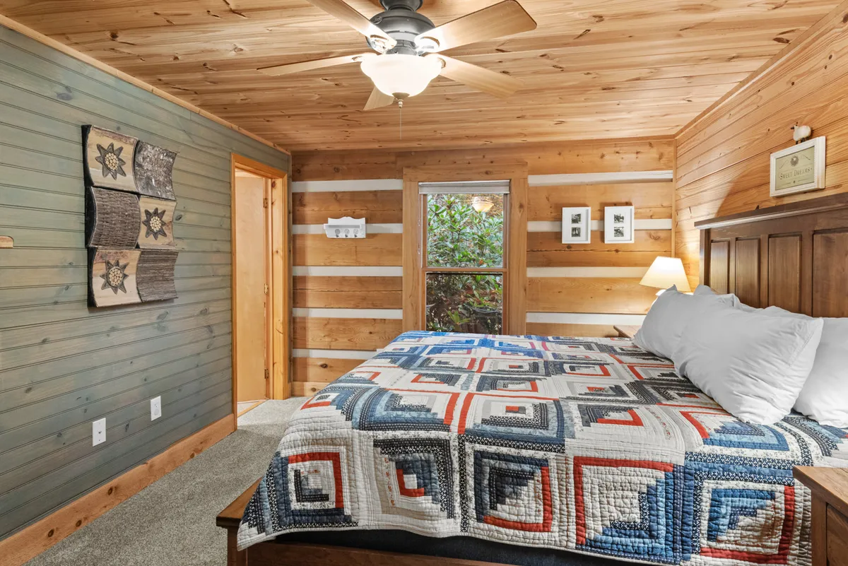 A comfortable bedroom featuring a quilted bedspread and a wooden headboard, positioned near a large window with views of the outdoors. Decorative touches and natural light enhance its tranquil, welcoming atmosphere.