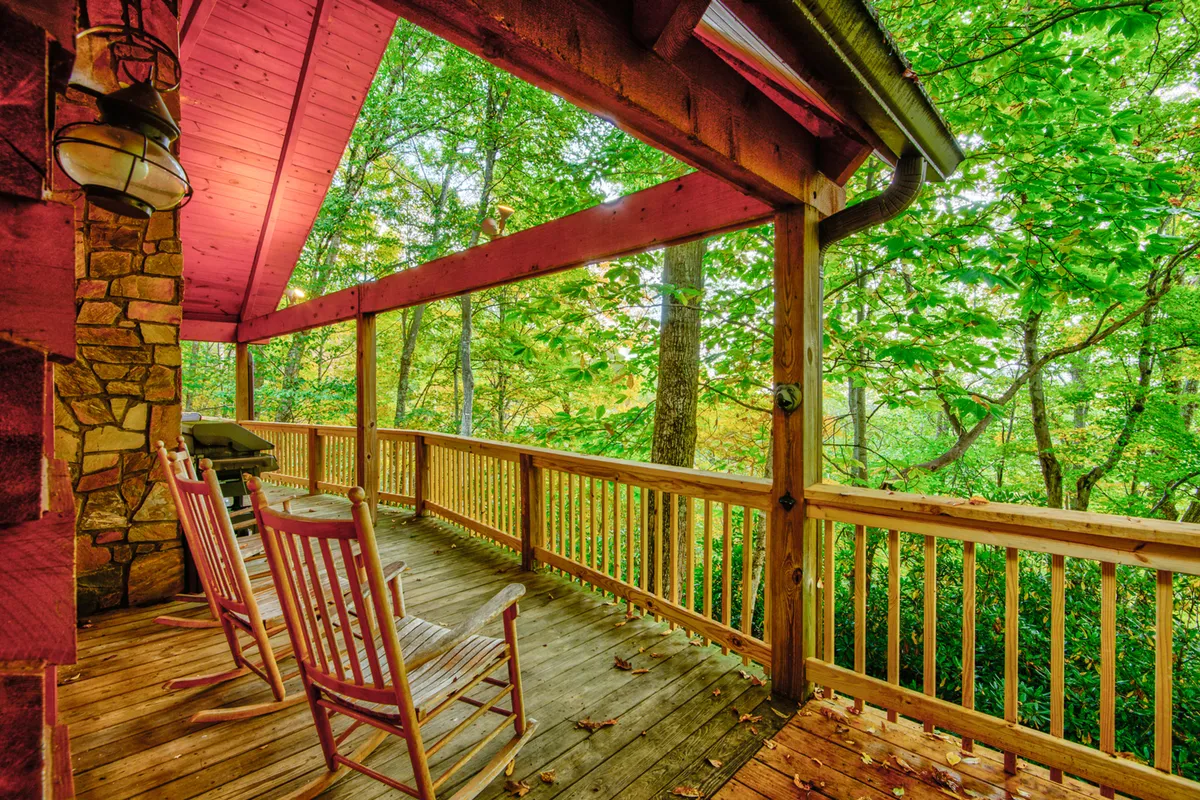 A peaceful covered deck featuring classic wooden rocking chairs set against a rustic stone wall, with lush green foliage creating a serene backdrop. The setting is ideal for relaxing at sunrise or sunset, with an inviting atmosphere for quiet conversations or tranquil reflection.