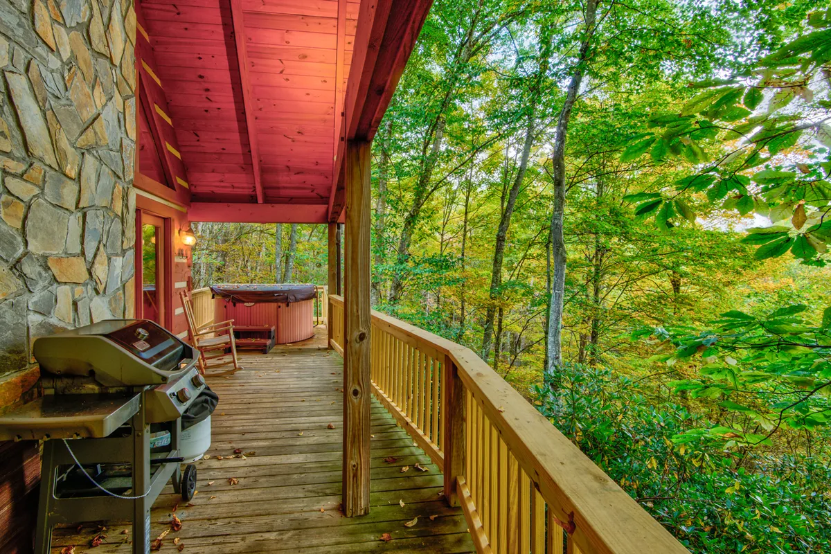 A wooden deck set among the trees, featuring a hot tub, a gas grill ready for barbecuing, and outdoor seating. The grill and the surrounding nature evoke the connection between indoor culinary experiences and outdoor mountain living.