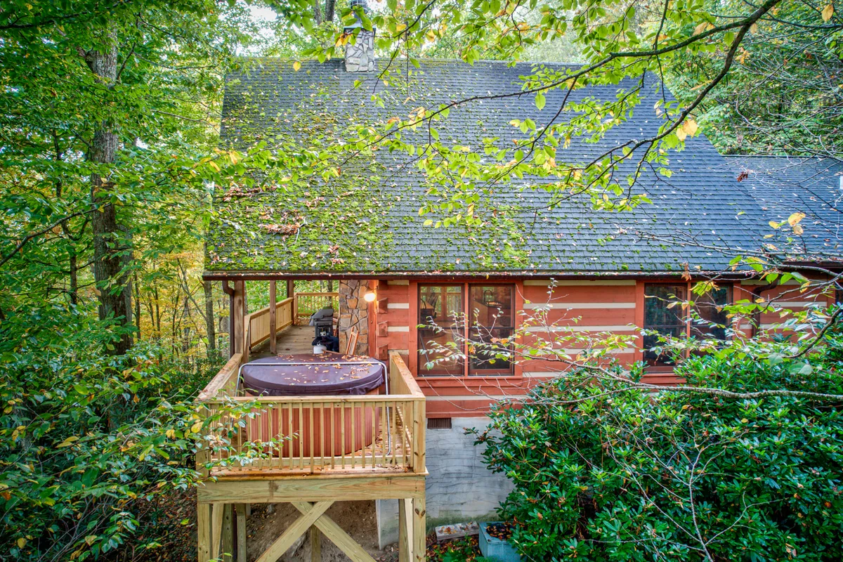 This image shows a tranquil log cabin nestled among lush greenery, with a hot tub prominently featured on the wooden deck. The surrounding trees create an intimate, private setting perfect for romance and relaxation. The deck invites guests to unwind while immersed in nature.
