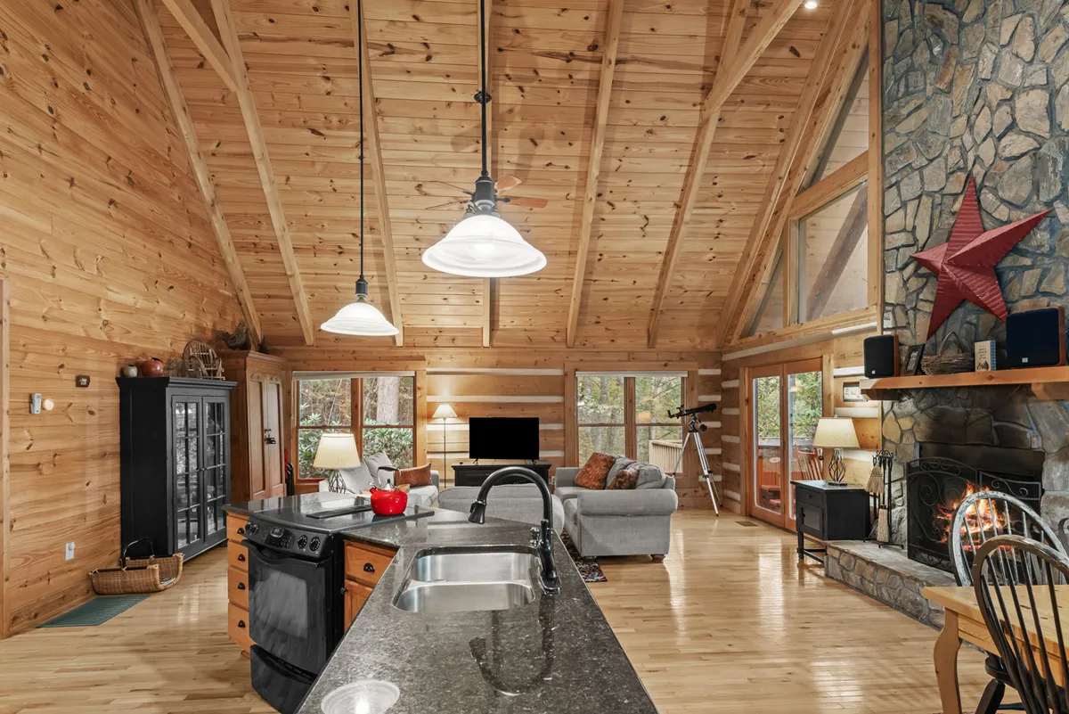 This image showcases an inviting open-plan living room that highlights the kitchen island, a wood-burning fireplace, expansive wooden beams overhead, and large windows providing panoramic views of the surrounding forest. The space is warm and welcoming, with multiple seating areas ideal for relaxing and entertaining.