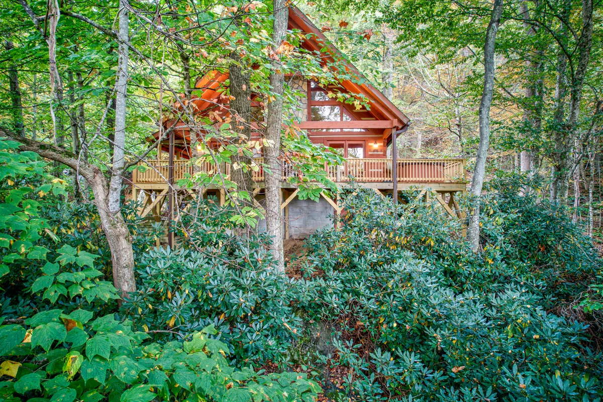 A serene exterior view of the cabin showcasing the spacious wooden deck bordered by large windows and surrounded by lush greenery and towering trees. The recognizable A-frame roof hints at a cozy, welcoming retreat ideal for groups.